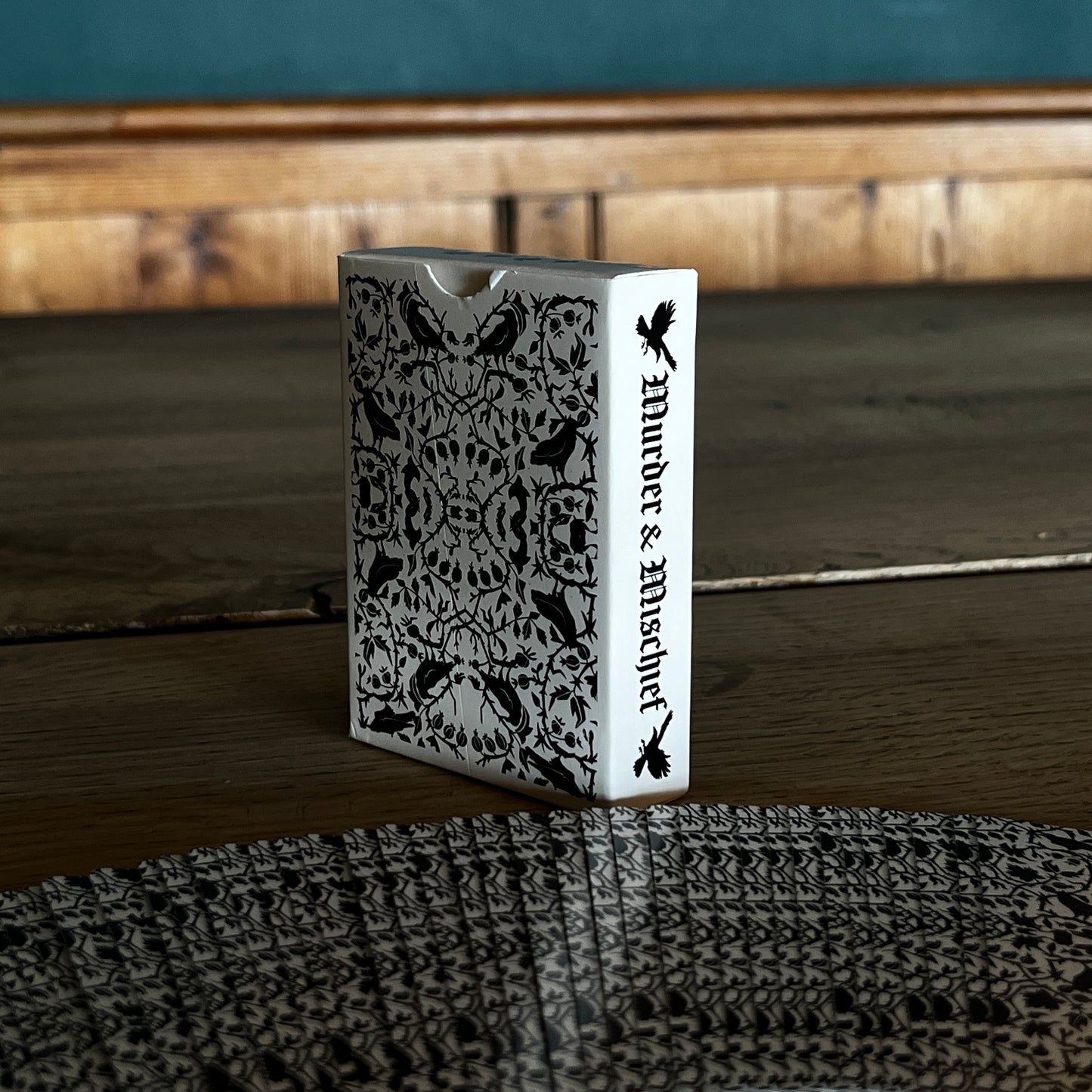 Corvid playing cards spread with the backs facing with the corvid and twined branches and leaf design, the spine of the box has Murder and Mischief in a gothic font.