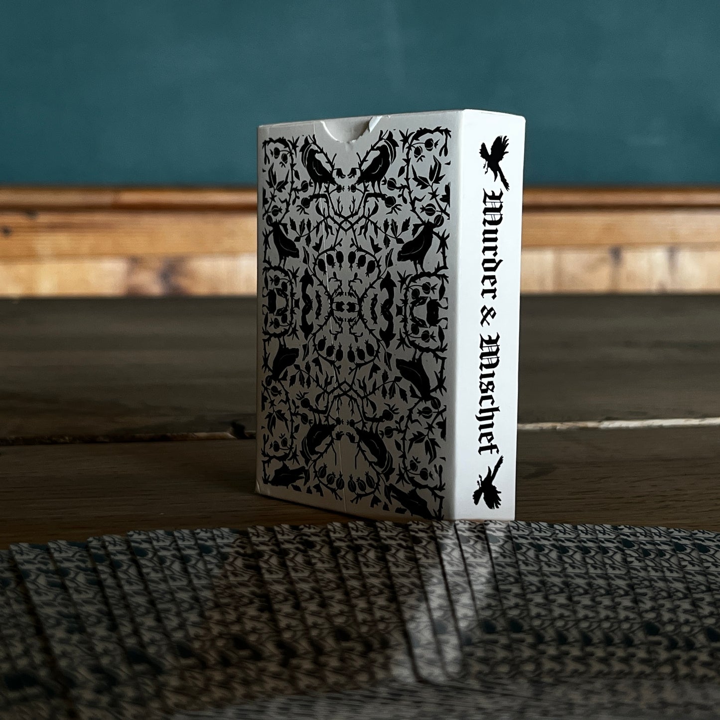 Corvid playing cards spread with the backs facing with the corvid and twined branches and leaf design, the spine of the box has Murder and Mischief in a gothic font.