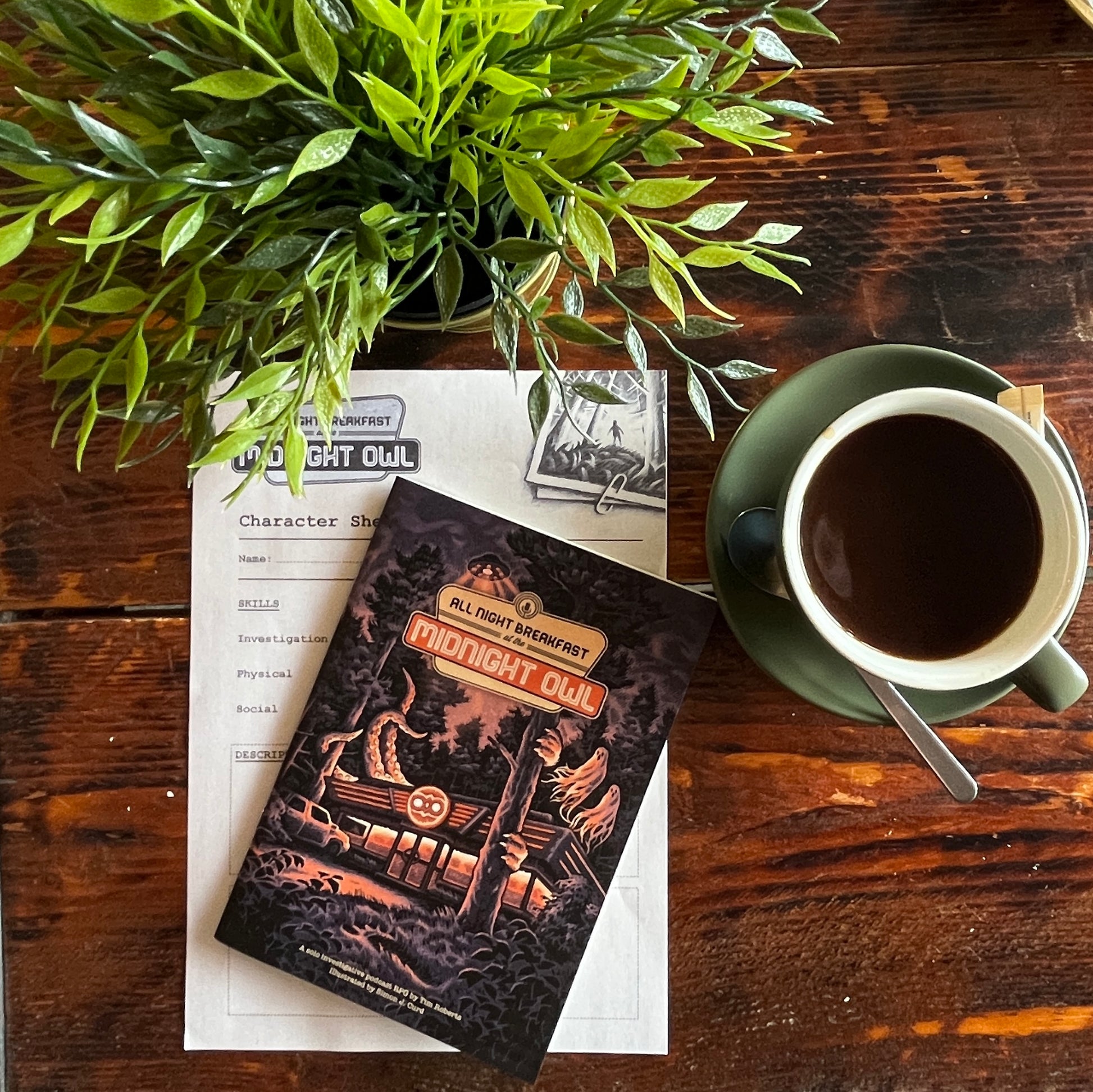 All Night Breakfast a cosy solo role playing game laid on a wooden table in a coffee shop with the character sheet, a cup of coffee and a leafy plant.