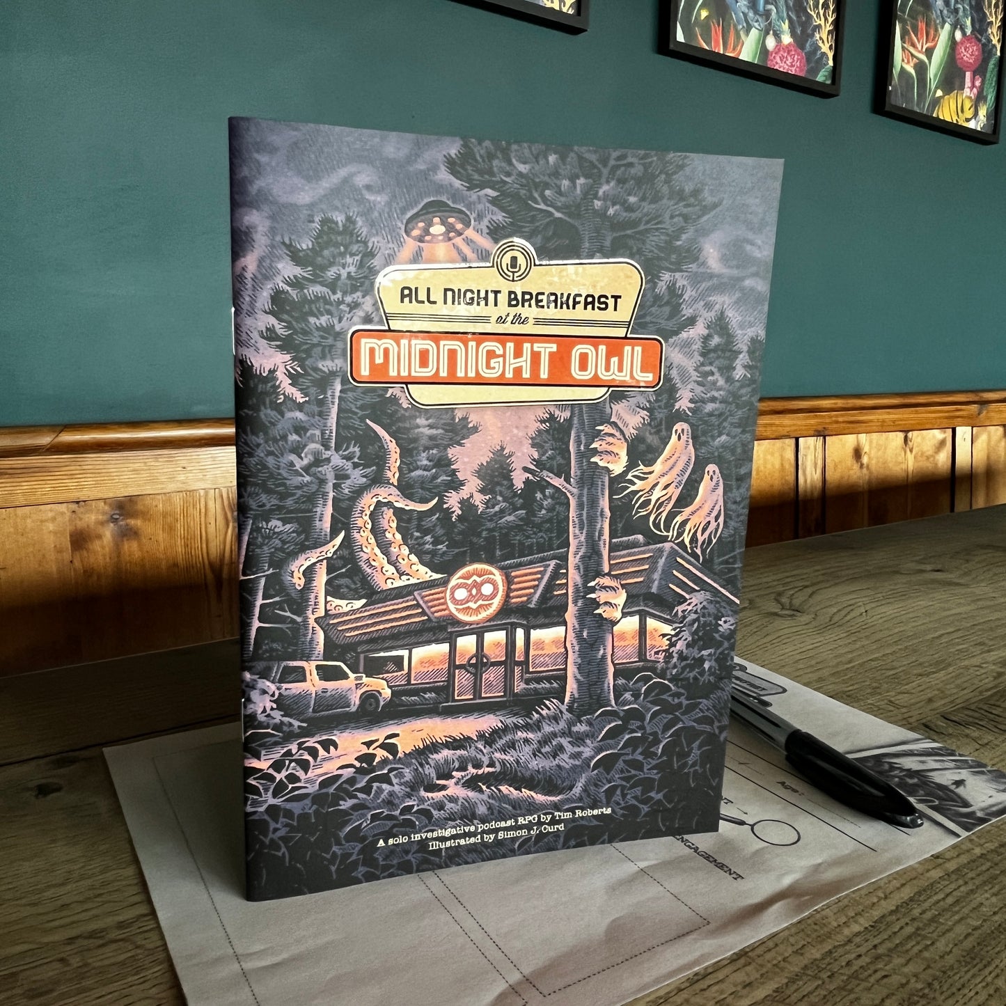 All Night Breakfast a cosy solo role playing game stood up on the character sheet with a pen in a coffee shop with a teal back ground