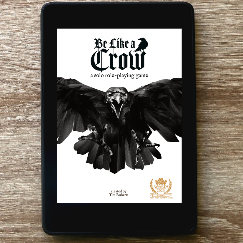 A black tablet device lying on a wood surface, displaying the cover art for a solo role-playing game titled "Be Like a Crow" with a large black raven graphic on a white screen.