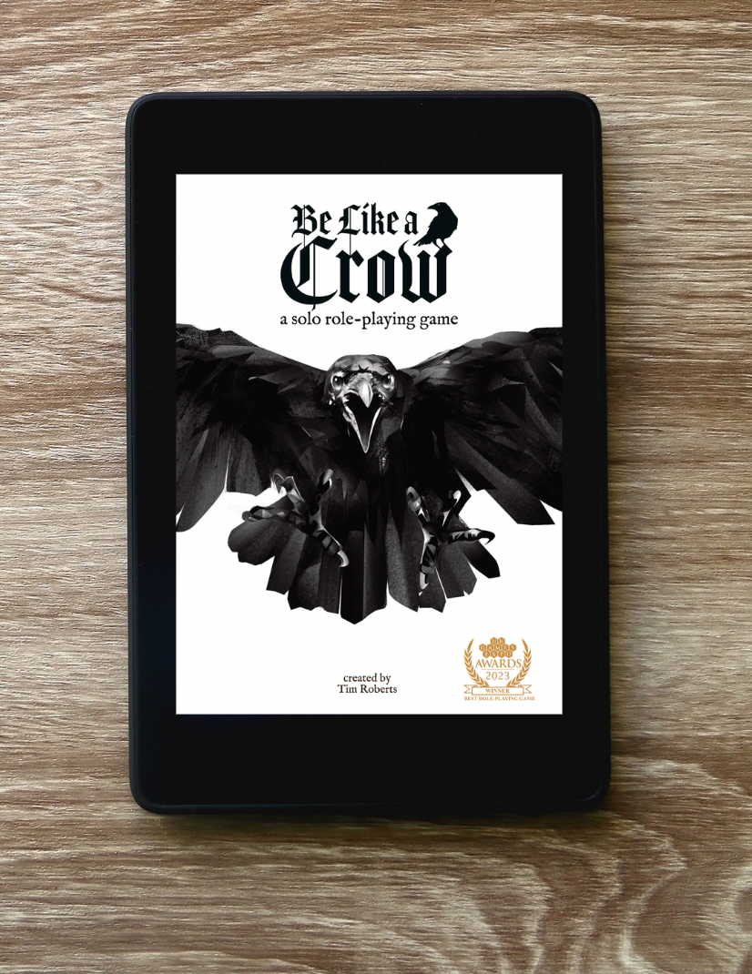A black tablet device lying on a wood surface, displaying the cover art for a solo role-playing game titled "Be Like a Crow" with a large black raven graphic on a white screen.