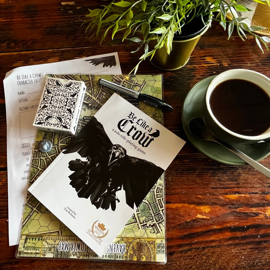Be Like A Crow coffee shop image with ravens of the tower pen, crow mini, corvid playing cards with a cup of coffee on a wood table.
