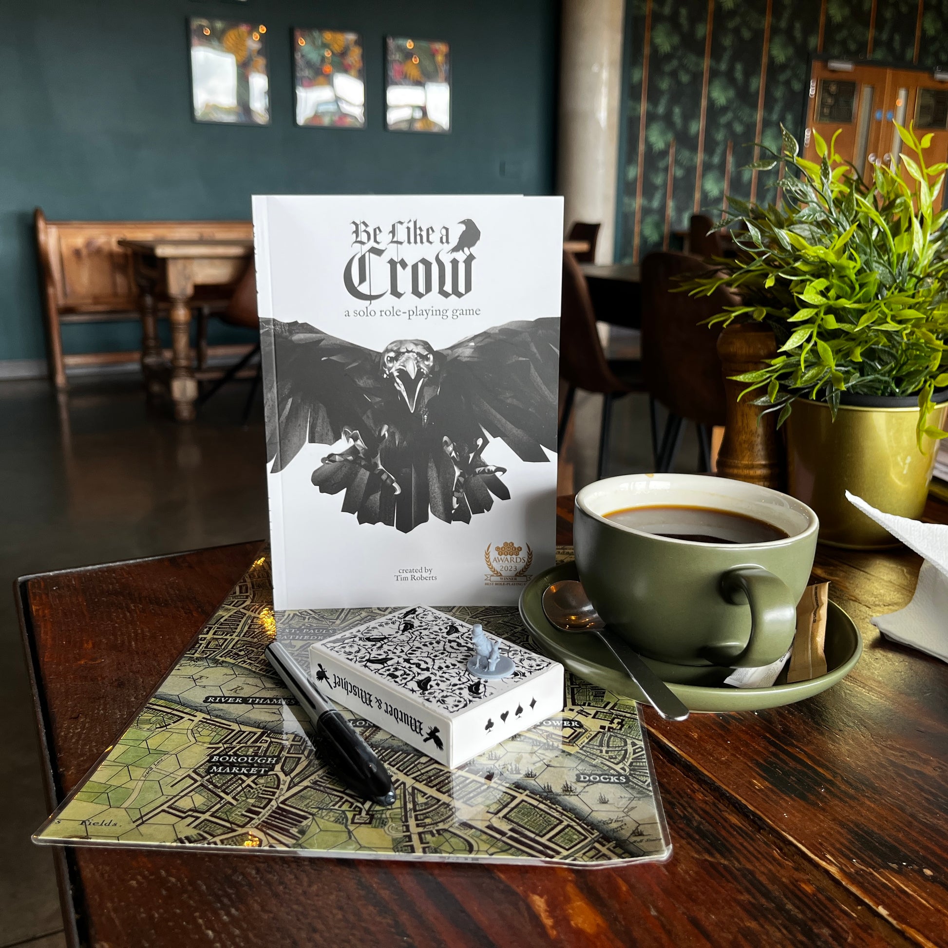 Be Like A Crow rulebook stood up in a coffee shop with the ravens of the tower map, crow mini, corvid playing cards, crow mini and a cup of coffee