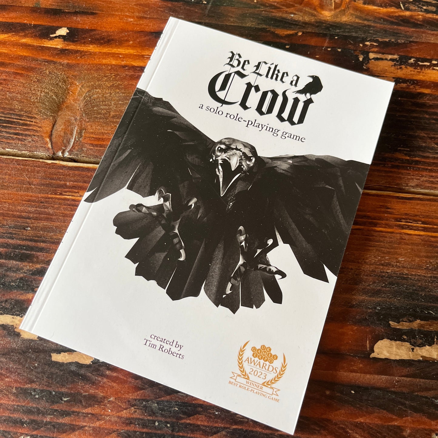 Be Like A Crow rulebook photographed on a rustic wooden bench, laid flat. The book as a large crow with open wings and the words Be Like A Crow in gothic font.