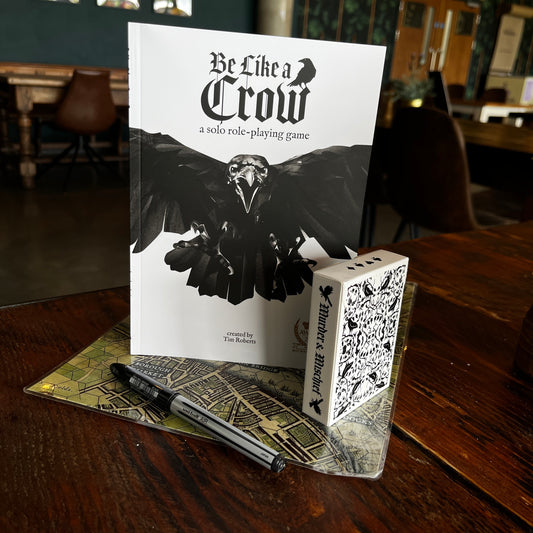 The Be like A Crow rulebook stood up with the corvid playing cards, on a map for Raven of the Tower and a pen set out in a coffee shop
