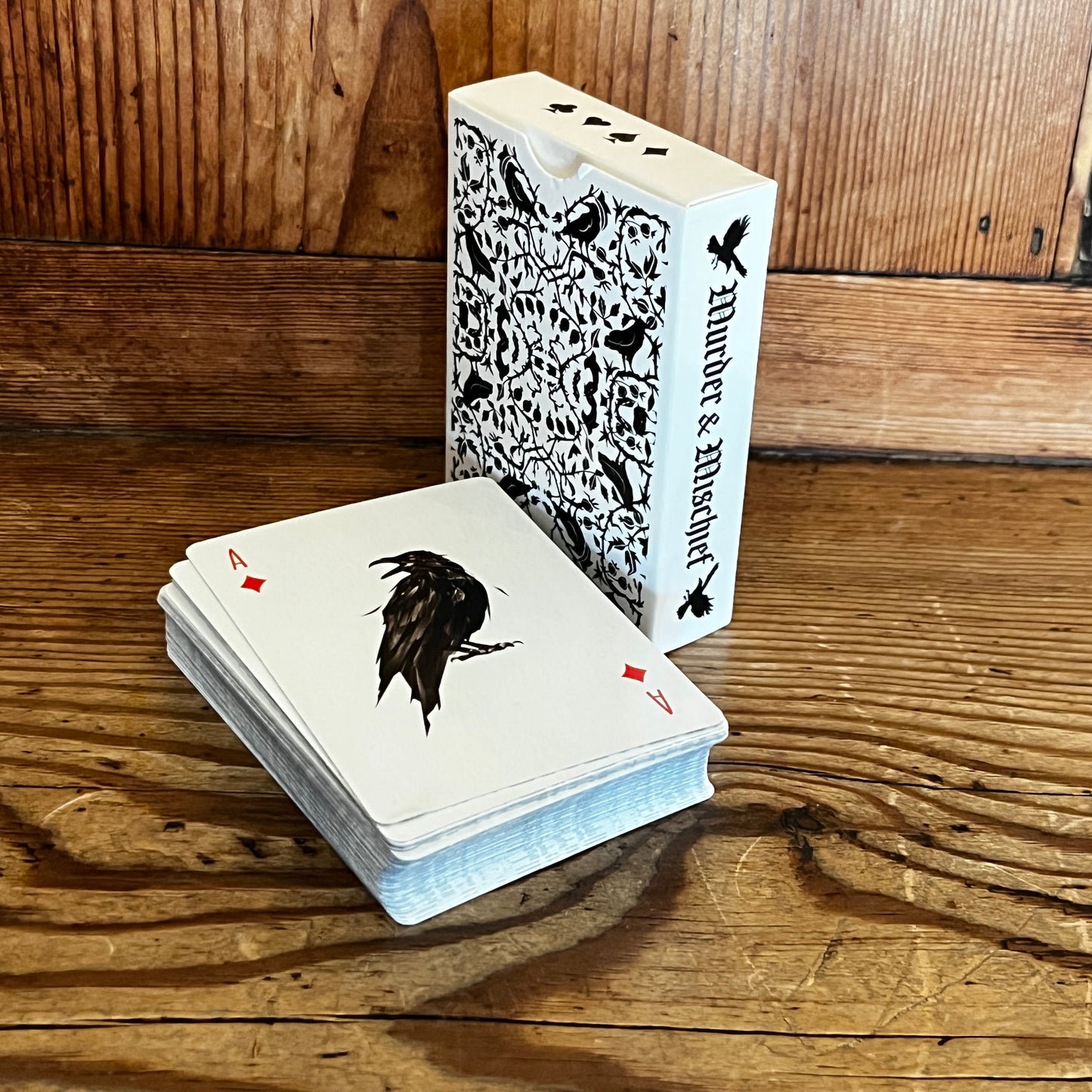 A deck of crow playing cards sat on a wooden bench with the box showing the spine with the title Murder and Mischief in a gothic font.