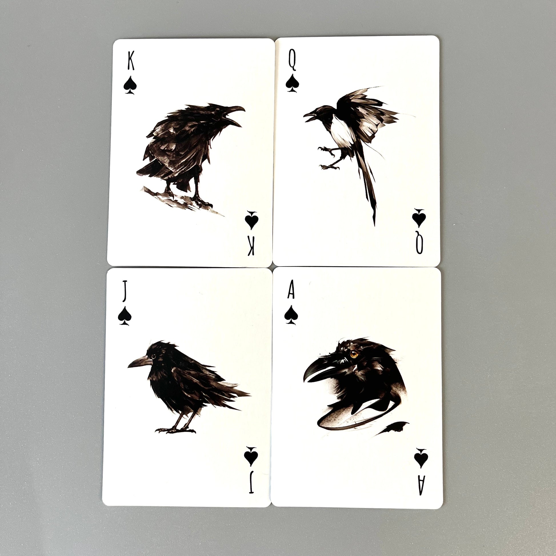 Four playing cards arranged in a 2x2 grid on a smooth gray surface. The cards are white with black illustrations of corvids and spade suit symbols, labeled K, Q, J, and A.