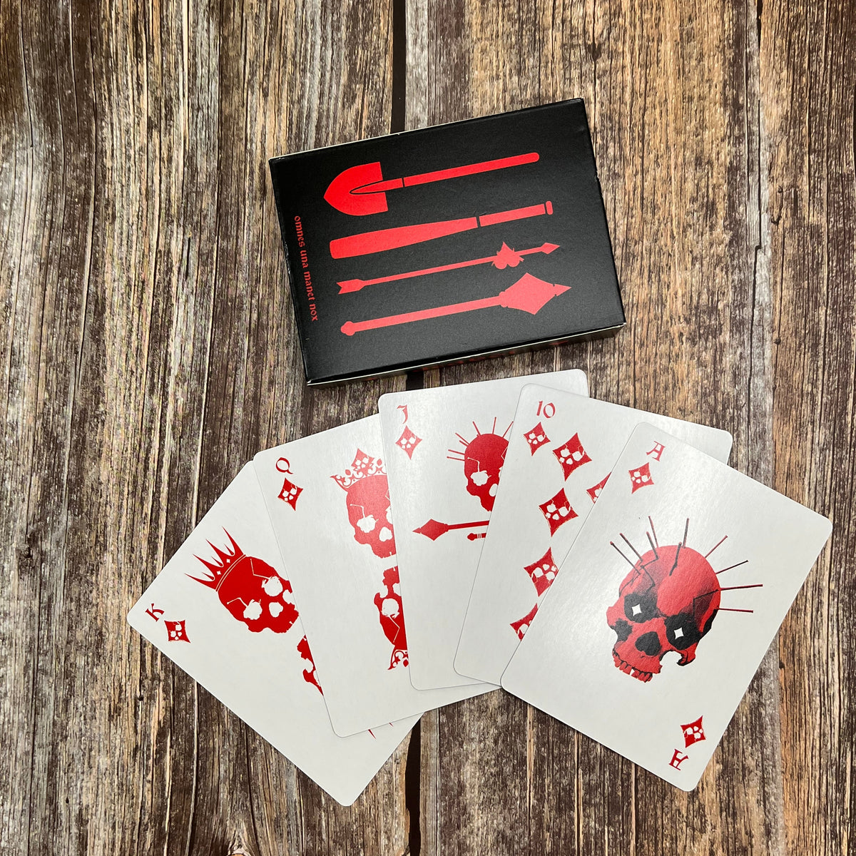 A set of red and white playing cards with skull and weapon-themed suits, arranged on a rustic wooden surface with their black and red box behind them.