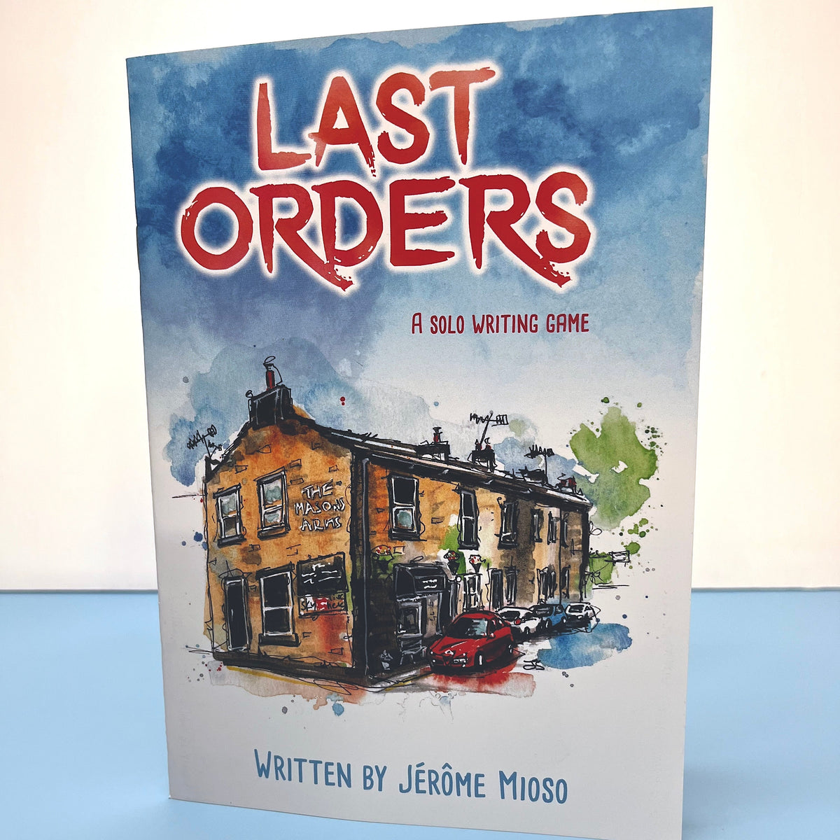 A single standing card or booklet titled "Last Orders" with a watercolor illustration of a row of terraced houses and the text "A Solo Writing Game" and "Written by Jérôme Mioso," photographed against a light neutral background with a blue surface below.