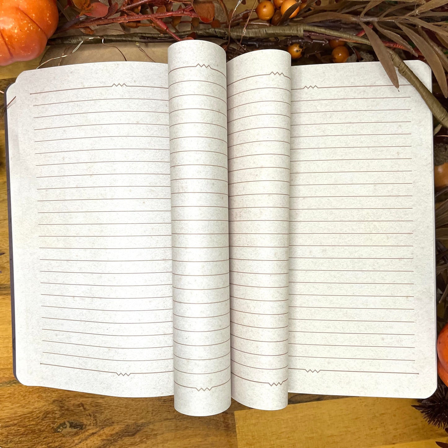 An open lined notebook or journal lies on a wooden surface, surrounded by autumn-themed decorative props including pinecones, dried leaves, and small pumpkins.
