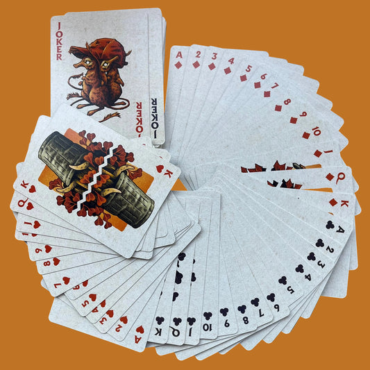 A deck of playing cards with several cards fanned out and a few face-up cards arranged on top, photographed on a smooth gray surface. The cards show illustrated suits and numbers, including a Joker card and 10s, 11s, and 12s visible in the spread.